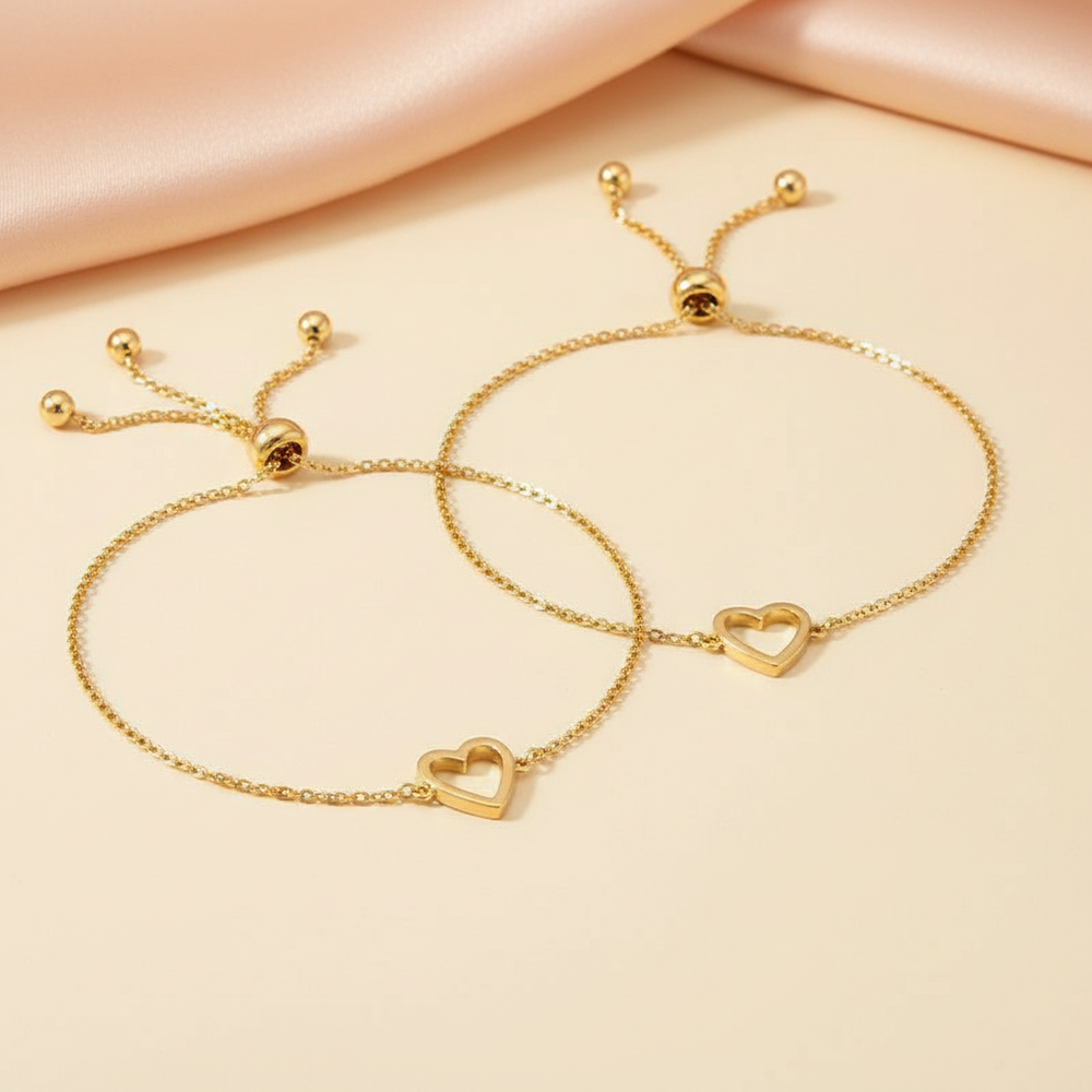 Three delicate 14K gold heart charm bracelets rest on a smooth, warm cream surface under soft golden light.