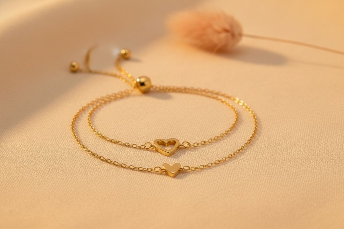 Three matching 14K gold heart charm bracelets displayed on a warm cream fabric surface with a soft golden glow.